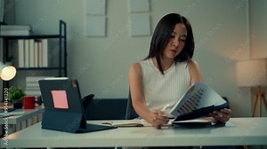 Businesswoman analyzes financial charts and takes notes at a modern office desk. Tablet and documents are neatly organized in a well-lit workspace