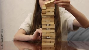 A woman carefully removes a wooden block from a tall tower of blocks, trying not to make the tower collapse