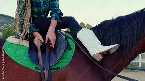 A cowgirl sitting on horseback secures the saddle. A young equestrian prepares for a horse ride