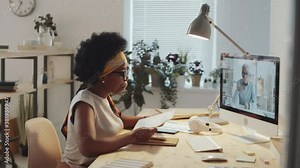 Young African American businesswoman sitting at office desk, having discussion with senior male colleague via online video call on computer and opening notepad for taking notes