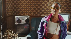 Stylishly dressed Black girl in 80s shell suit moving her head to the beat and enjoying the music in old room, analog TVs with no signal screen and boombox in the background