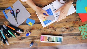 Zoom-out of young woman drawing with crayons in notebook making original collage sitting at desk at workplace indoors. Art, people and imagination concept.