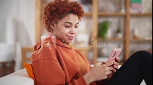 Beautiful happy young african american girl sitting on sofa in living room at home using mobile phone. Online communication distance friends online in mobile chat app using smartphone video chat.