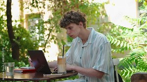 Young man teenage student studying, working on laptop at cafe outdoors. Male student learning using computer, making notes. Distance education concept, freelancer, work remotely in summer