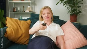 Blonde woman watching television, while sitting on sofa in living room. Pensioner looking for tv show program, using remote controller. Spare time, pension at home, self-isolation during covid.