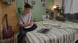 Full shot of teenage Caucasian male student sitting in bedroom, with cochlear implant connected to laptop, listening to online college lecture and writing in notebook, while studying for exam