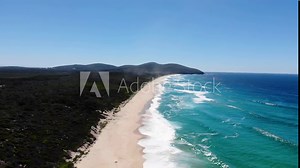 Drone flying above beach on sunny day with blue skies with aerial views of ocean waves and water crashing on to sandy beach from high angle