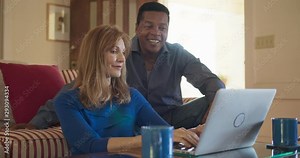 Older African American and Caucasian husband and wife using laptop computer together in living room. Mature homeowner married couple smiling while planning trip online. Slow motion 4k handheld