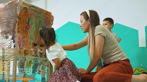 Cling film painting. Toddler painting with a sponge, brushes and paints on a cling film wrapped all the way round the wooden shelf unit. A teacher helping them. Creative activity for kids sensory