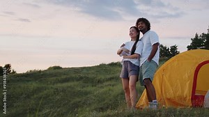 Young couple in love having a great time in the mountains by pitching a tent and enjoying a summer evening hugging, communicating and watching the sunset.