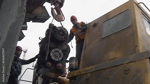 Engineering Specialists Assisting Industrial Crane Lifting Machine Engine On Hook. Specialists Control Process Of Machine Engine Relocation. Specialists Inspecting Heavy Machine Engine. Maintenance