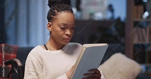 Young pretty african serious woman sitting in wheelchair browsing internet news on mobile tablet computer digital device at home.