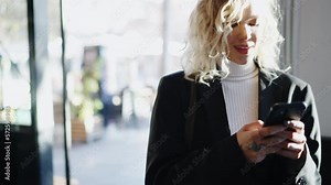 Pretty blonde hair female enters an cafe with cellular phone in hands and reads incoming text messages. Smiling young woman chatting online through cellphone app. People using technology. Slow motion