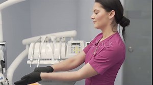 Young Female Dentist in Purple Uniform Putting Gloves on Her Hands and Mask on Her Mouth and Looks at Camera. Some Medical Instruments in Background