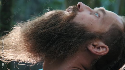 A close-up shot of a young man with a long beard and hair bun performing yoga stretches in nature. Focusing on his neck and face as he transitions between poses with sunlight filtering through the tre