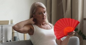 Stressed middle aged older woman sitting on sofa, using paper fan. Overheated sweaty mature female retiree suffering from hot summer weather high temperature inside without air conditioner at home.