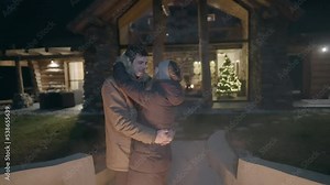 Slow motion - A young brunette couple, dressed in winter clothes, slow dancing and kissing outside a modern cottage, making eye contact, on a cold winter night.
