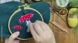 Women's hands embroidery leg of mushroom with floss threads on tambour. Sewing of fly agaric on green beret. Handicrafts workshop. Wooden table with yarn scissors and candle on background.