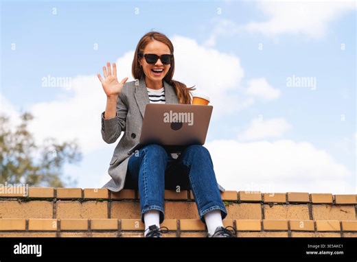 Friendly young woman waving hello during a video call on a laptop while sitting on urban steps outdoors Stock Photo - Alamy