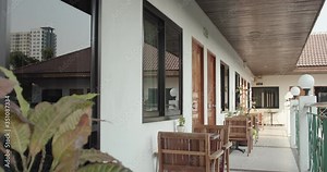 Exterior hotel corridor in the tropical sunny day, wooden tables and chairs in front of guest rooms. Hotel corridor is decorated with small tropical plants. Residential building, hotel, accommodation.