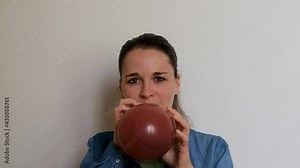 Girl is preparing for holiday and blowing balloon and then lets it fly away. Young pretty woman of Caucasian European appearance inflates burgundy balloon with her mouth on white background.