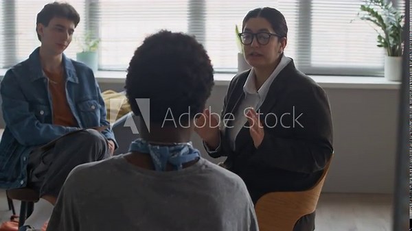 Young confident biracial female school counselor speaking to group of diverse teen students sitting on chairs in circle in cozy office listening to her