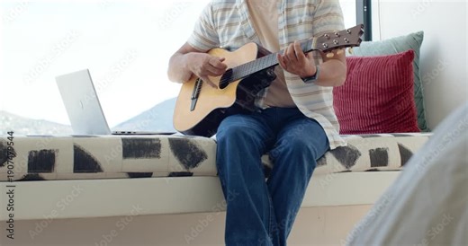 Man is switching chords and plucking guitar on window bench, following laptop backing track
