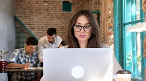 Young Attractive Woman Working on her Computer. Looking at Display. Wearing Eyeglasses.Surfing the Internet. Typing Mail Messages.Working on Start up. Group of Business People in the Background.