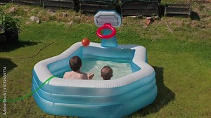 Two boys sitting in inflatable pool, playing with a basketball and aiming for inflatable basketball hoop on sunny day.