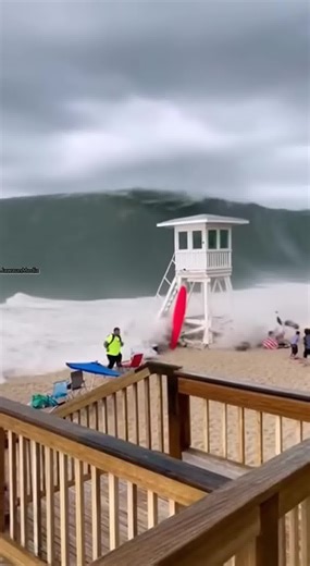 Massive Wave Destroys Lifeguard Tower, Floods Boardwalk 🌊⚠️ Ocean City, New Jersey — Tuesday, February 25, 2025 Vertical smartphone footage filmed from a raised wooden boardwalk shows a towering dark-green wave already slamming into the shoreline as the video begins. The surge strikes a white wooden lifeguard tower, snapping its stilts and collapsing the structure instantly into the water. The wave continues rushing forward at high speed, sweeping up rows of colorful beach umbrellas and coolers