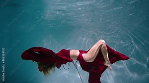 Magical underwater shot of a young woman swimming underwater. Red dress and woman's hair fluttering in blue water column with glare. Subaquatic shot in pool, close up in slow motion. Stock Video