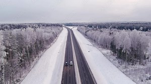 Aerial shot of a four lane highway in Estonia during wintertime. Cloudy sky. Snowy forest and ground. Cold nordic weather. Vehicles driving. Drone slowly zooming out.