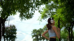 Side view of young beautiful asian woman jogging outdoor at village park, sporty female running in the garden, cheerful girl enjoy morning fitness while listening music via headphone.