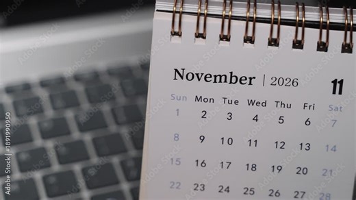 Close-up of a desk calendar on a laptop keyboard as a hand flips the page from October 2026 to November 2026. Camera stays static, showing a clear monthly time transition and structured planning conc