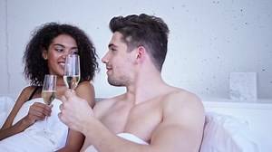Smiling loving multiethnic couple drinking champagne and talking while lying in bed
