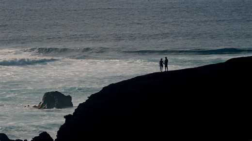 A young couple walks hand in hand along a rugged volcanic cliff overlooking the Atlantic Ocean in Lanzarote, Canary Islands, Spain. | Premium Stock Video Footage