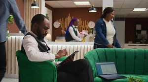 African american staff making online bookings on tablet in lobby, working at luxurious hotel and providing excellent concierge service for guests upon their arrival. Bellboy in uniform manage rooms.