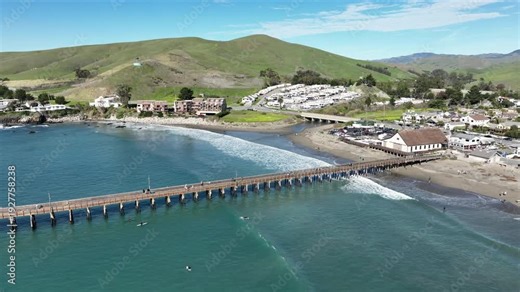 Aerial view of Cayucos, California USA featuring Cayucos Pier in downtown, Pacific Ocean waves, tourists along Ocean Ave, grass fields and Highway 1 on the scenic Central Coast in San Luis Obispo
