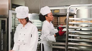 Two professional female chefs in white cook uniforms and aprons knead pastry dough and eggs, prepare bread, cookies, and fresh bakery food, baking in oven at a stainless steel kitchen of a restaurant.