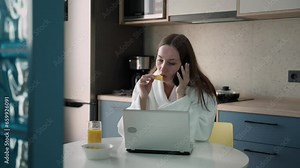 Beautiful woman in a bathrobe uses a laptop and talks on a mobile phone while spending time in the kitchen.