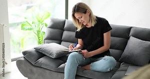 Smiling woman makes notes in diary sitting on cozy sofa at home. Inspired blonde female engages in planning routine creating schedule