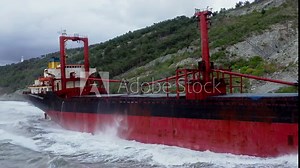Sea waves are breaking on big red cargo ship that is aground. Freighter went ashore. Bulk carrier got upon bank. Watercraft is on seashore near mountains. Barge was stranded. Navigation, shipping