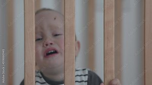 portrait of crying tears screaming baby boy child in cradle crib cot. caucasian toddler kid face behind wooden grid bars. baby in striped clothes has scratch scar on forehead. bars in hands