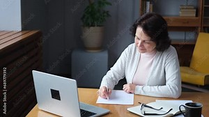 Middle aged senior woman using laptop computer writing notes at home. Focused mature old businesswoman work in home office making written records doing online work. Mature boss professional worker