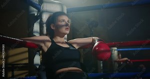 Close-up of tired female boxer resting on boxing ring ropes after intensive training. Exhausted athlete breathing hard overcoming failure during competition.