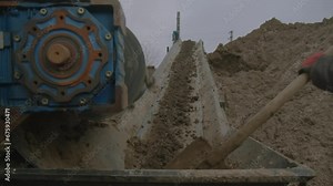 Worker removes dirt from under rotating roller on clay transporting conveyor closeup. Employee with shovel controls soil carrying in quarry