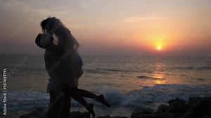Bride silhouette kissing groom holding woman in arms enjoying romantic twilight. Married couple embraces kissing against ocean at sunset
