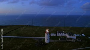 Aerial orbit around the illuminated Loop Head lighthouse, revealing the Atlantic Ocean at dawn.
