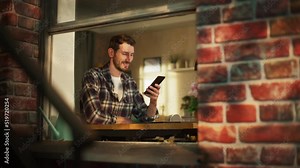 Happy Smiling Man Sitting Behind Desk at Home Office and Using Smartphone. Handsome Man Browsing Through Internet, Checking Videos on Social Media, Shopping Online. View From Outdoors into Window.