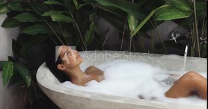 Young woman blowing soap bubbles and having fun while lying in a bathtub full of foam. Tropical home spa concept. Charming lady relaxing in the bath and enjoying a beauty and skincare day